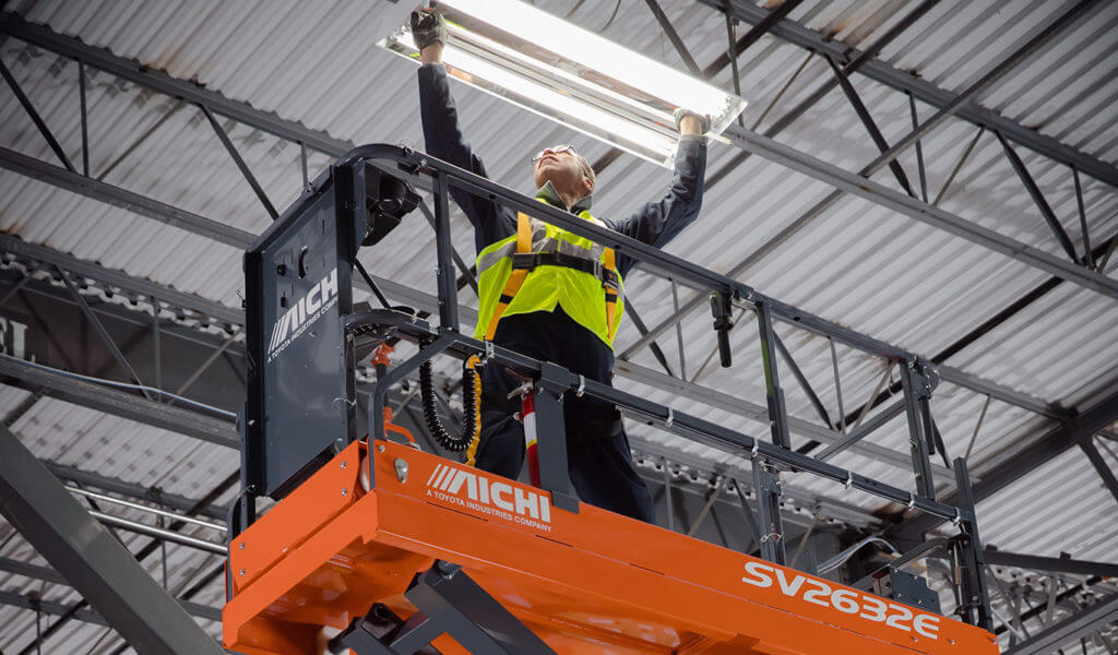 A worker uses a scissor lift to change out a lightbulb installed in the ceiling of a warehouse