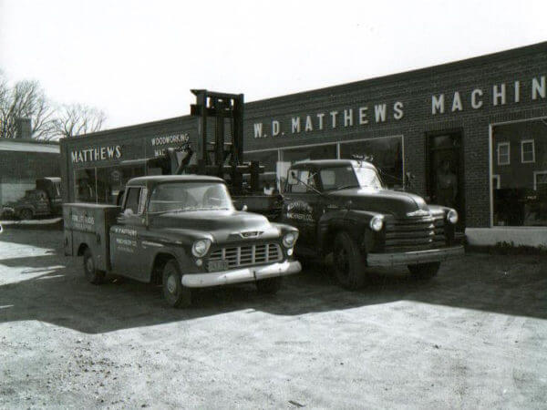 A vintage photo showing two old service trucks parked outside of the WD Matthews building