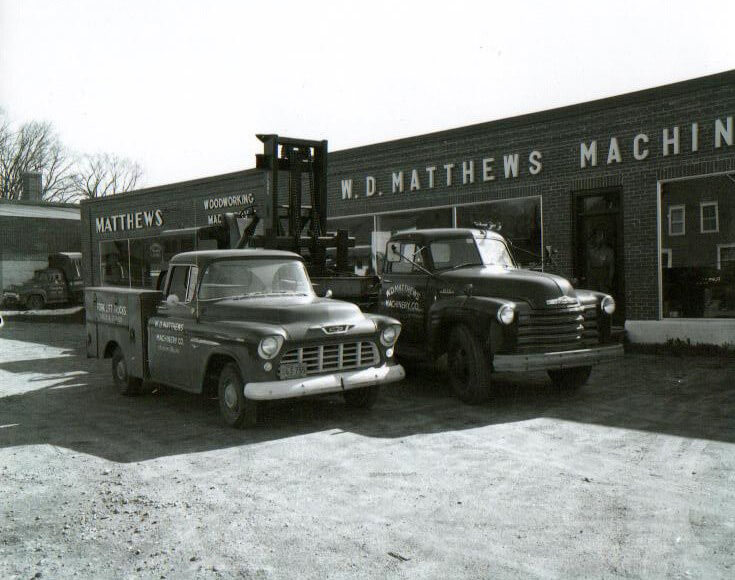 A vintage photo showing two old service trucks parked outside of the WD Matthews building