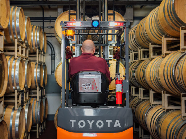 A man uses a Toyota forklift to navigate through a narrow aisle of wine barrels stacked on top of each other