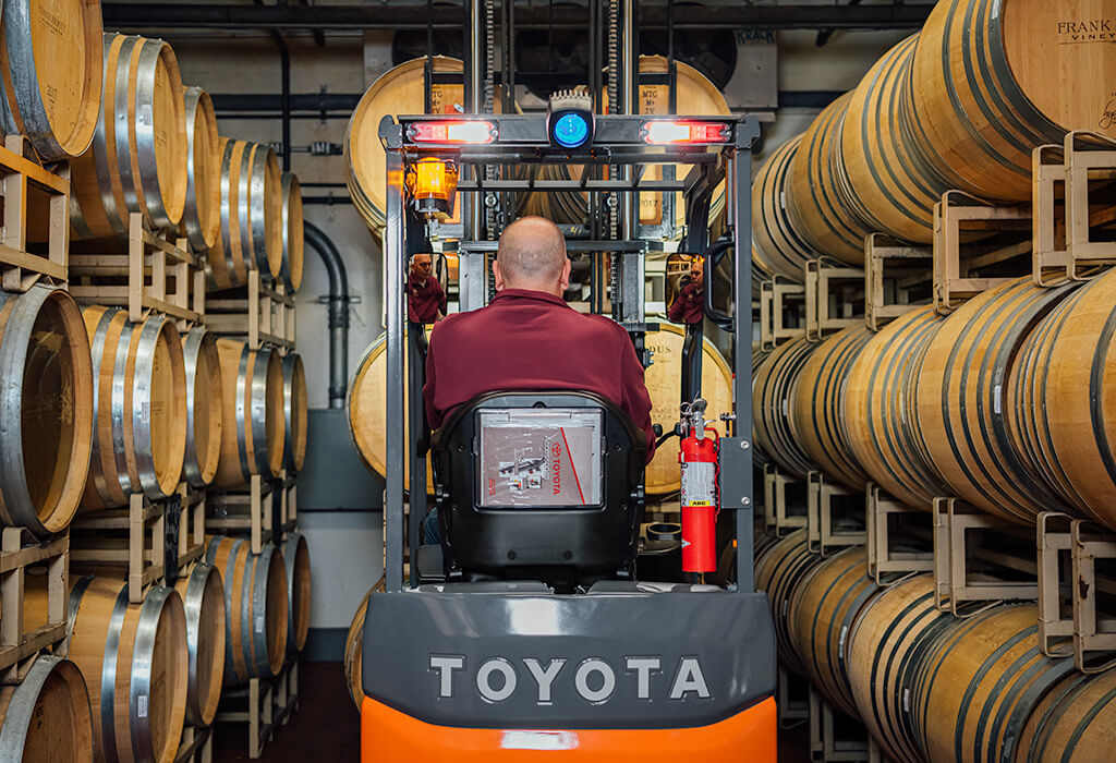 A man uses a Toyota forklift to navigate through a narrow aisle of wine barrels stacked on top of each other