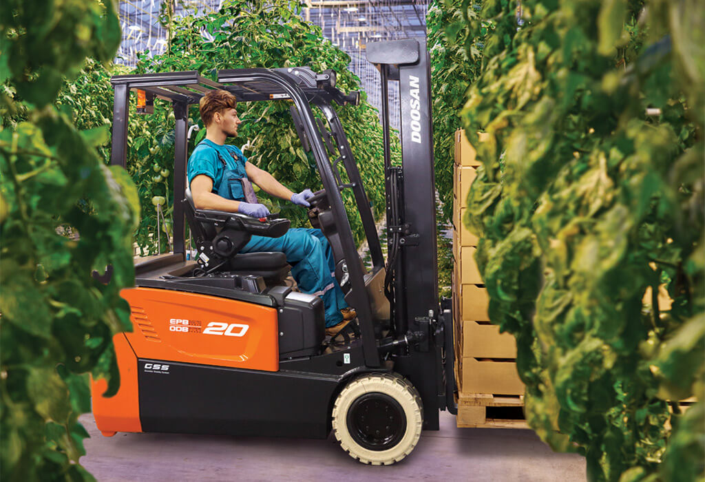 A young worker maneuvers a forklift through narrow aisles of large, leafy plants growing in a greenhouse