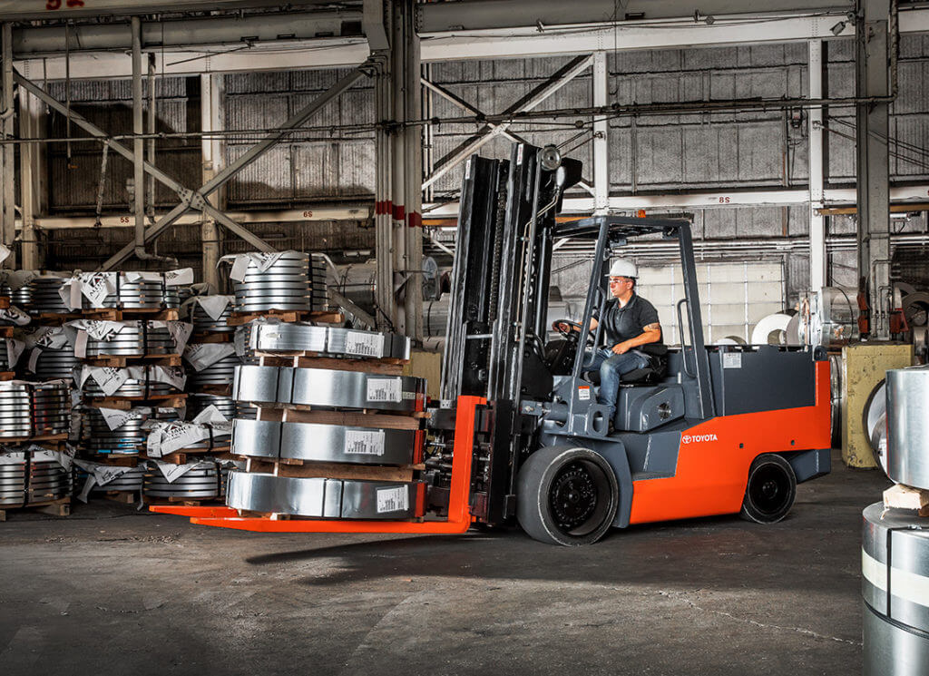 A worker uses a forklift to transport heavy metal sheeting in a warehouse setting
