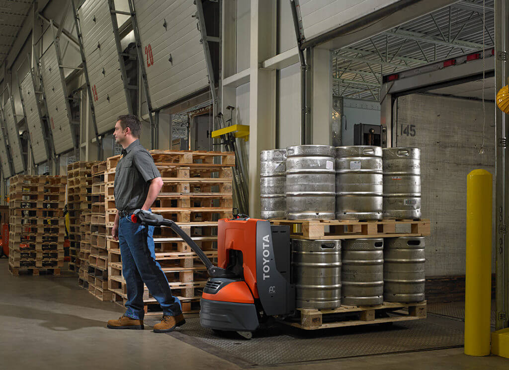 A man uses a hand pallet truck to pull heavy metal kegs in a warehouse setting