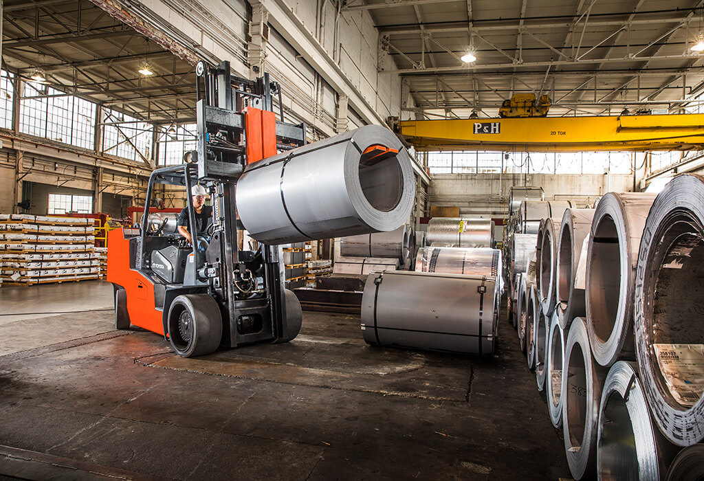 A worker uses a forklift to lift a heavy piece of rolled metal sheeting