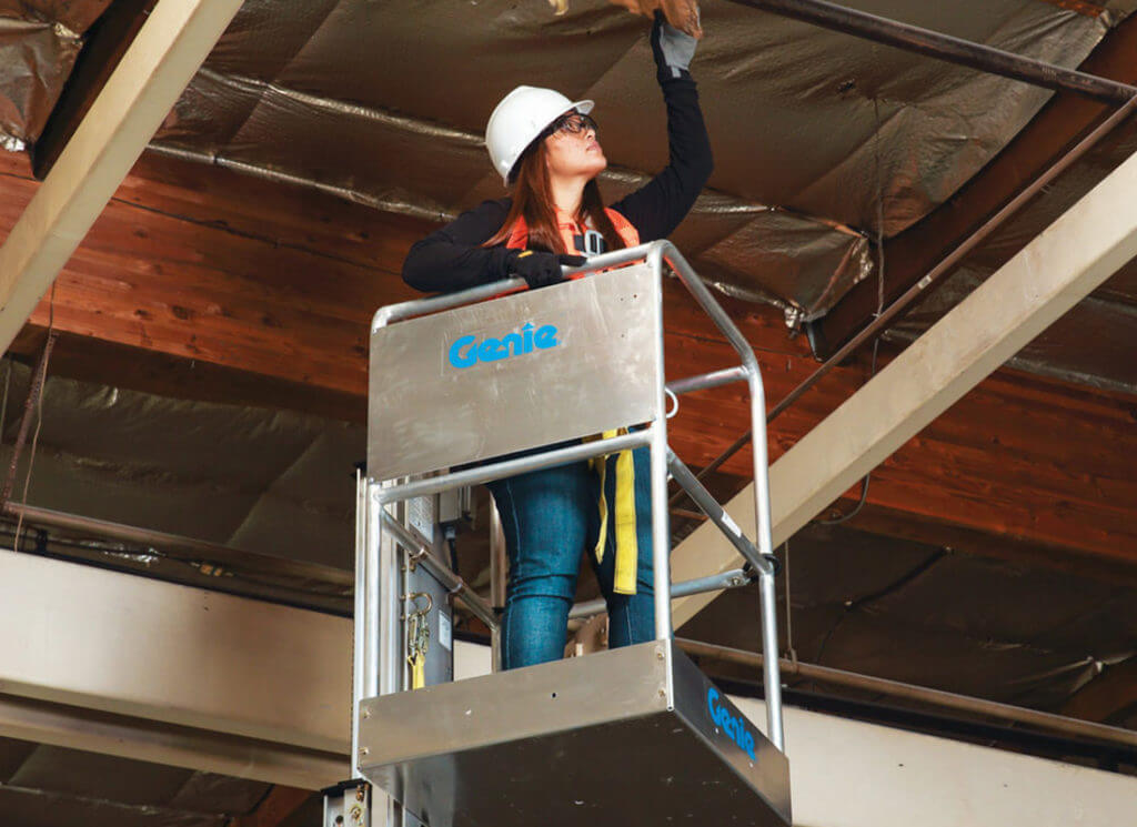 A woman stands on an aerial lift to pull insulation from a ceiling in an industrial setting