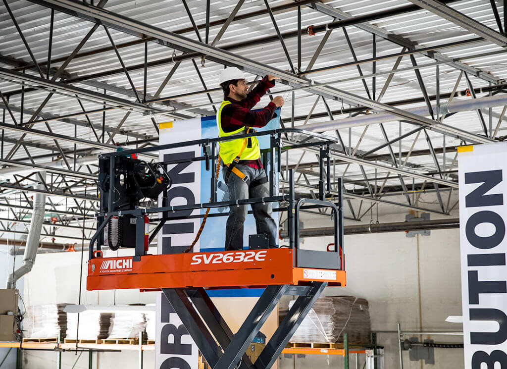A man uses a scissor lift to attach a long hanging banner to a ceiling in preparation of a tradeshow